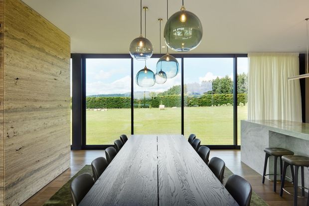 A refined contemporary dining area with a long dark timber dining table and black upholstered chairs, positioned beside full-height black-framed glazing overlooking a broad lawn and landscaped garden. A cluster of suspended spherical glass pendant lights hangs above the table. A pale textured stone or timber-look feature wall stands to the left, while a stone kitchen island with stools is visible on the right behind a sheer curtain.