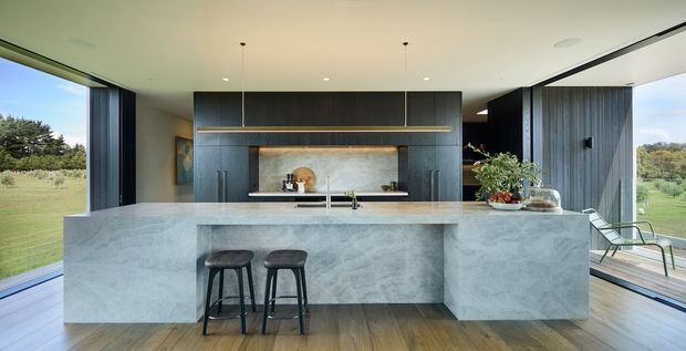 A refined contemporary kitchen with a large pale stone island and waterfall ends, two dark stools, dark vertical-grain cabinetry, and a full-width integrated rangehood and cooking wall. Warm timber flooring contrasts with the cool grey stone and cabinetry. Expansive corner glazing opens the kitchen to a green landscape, creating a bright, open setting.