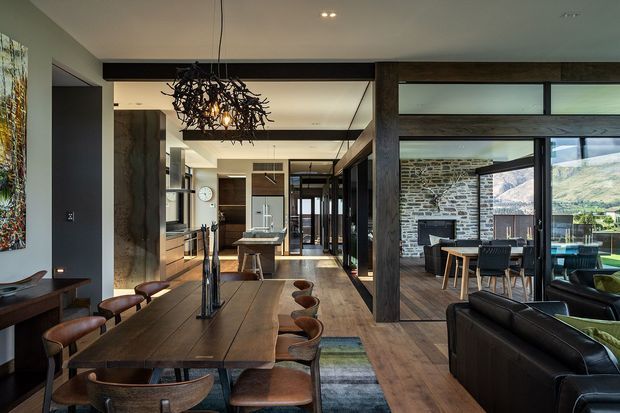 Open-plan contemporary interior combining a dining area, kitchen and living space. A long dark timber dining table with leather chairs occupies the foreground, while a kitchen with island and built-in cabinetry is visible beyond. The living area has dark leather seating and opens through extensive black-framed glazing to a stone-walled area and mountain landscape. Exposed dark timber beams, timber flooring, recessed lighting and a sculptural branch-like pendant create a refined, warm modern character.