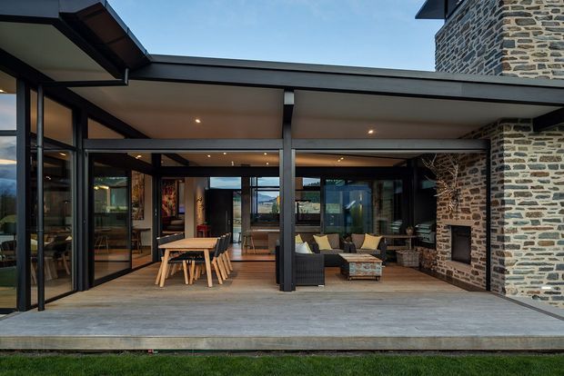 Exterior view of a contemporary covered patio and outdoor living area, with extensive floor-to-ceiling glazing, dark steel framing, a broad flat roof, timber decking, dining furniture, lounge seating, and a prominent rough stone fireplace chimney wall.