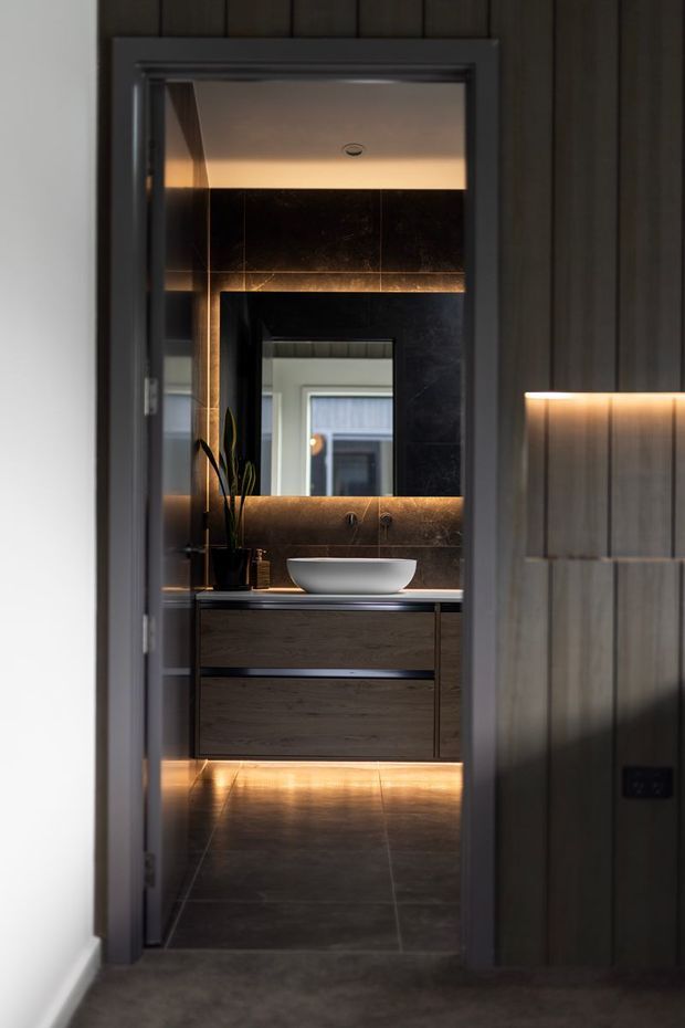 A dark, refined bathroom viewed through an open doorway. The room features large dark stone-look wall and floor tiles, a floating dark timber vanity with integrated drawers, a white vessel basin, wall-mounted taps, a large backlit mirror, and warm concealed LED lighting beneath the vanity and along the mirror. Vertical dark wall panelling and a softly illuminated built-in niche are visible in the foreground.