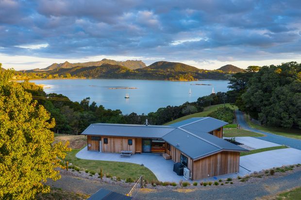Aerial exterior view of a low-profile contemporary timber-clad house with multiple pitched metal roof planes, set on a landscaped sloping site overlooking a sheltered bay with sailboats and rugged hills. The building includes a broad paved driveway and paths, established trees, and a courtyard-like area around the main structure.