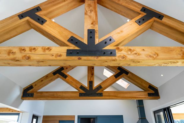 Interior view looking up at exposed timber roof trusses with prominent black metal connector plates and bolts. The warm wood structure spans a bright, contemporary room with white walls, skylights, and dark-framed glazing visible below.