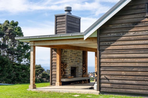 Exterior view of a contemporary timber-clad house with a steep gabled roof and a covered outdoor living area. The shelter contains a substantial exposed-brick and stone fireplace, with timber posts and decking or concrete paving. A chimney rises above the roof, and a distant coastal landscape is visible beyond the structure.