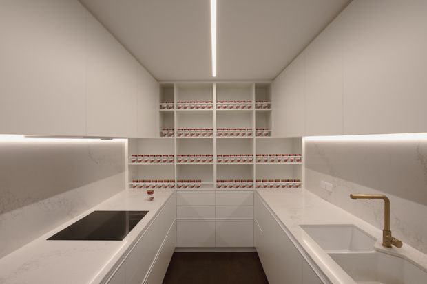 Compact contemporary galley kitchen with white built-in cabinetry, pale stone benchtops and integrated backsplash surfaces. Open shelving at the rear is filled with neatly arranged jars, while a black induction cooktop sits on the left and a double-bowl sink with a brass-toned tap is on the right. Continuous concealed LED lighting runs beneath the upper cabinetry and along the ceiling, creating a clean, softly illuminated space.