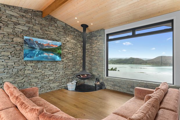 A cosy living room with a large corner window overlooking a lake and hills. The room features exposed stacked-stone walls, a warm timber-lined ceiling with exposed beam, wide timber flooring, a suspended black wood-burning fireplace, and a curved peach-toned sectional sofa arranged around the fireplace and wall-mounted television.