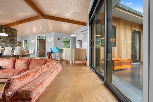 Open-plan living and kitchen interior with a large blush-toned sectional sofa, timber flooring, white walls, exposed warm timber beams and a pale timber-lined vaulted ceiling. A stone-clad feature wall and fireplace are visible at left, while the kitchen has light cabinetry and a timber island. Full-height black-framed glazing opens to a covered outdoor room with timber wall cladding and built-in bench seating.