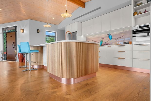 Bright contemporary kitchen with a large curved island clad in vertical timber slats and a metallic-toned plinth. The space features white full-height cabinetry, integrated appliances, a colourful bird-themed splashback, timber flooring, and a warm timber-lined ceiling with exposed beam detailing. A breakfast bar with turquoise stools sits beside the island.