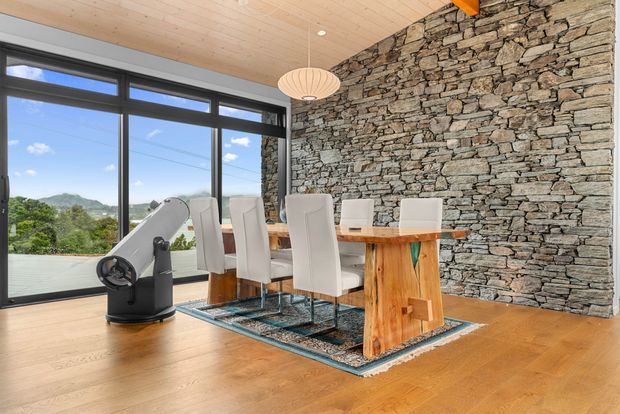 A bright dining area with a long rustic timber dining table and white upholstered chairs, set against a dramatic dry-stacked natural stone feature wall. Full-height dark-framed glazing opens to a green mountainous landscape, while a light timber-lined ceiling, warm timber floor, pendant light, and telescope complete the room.