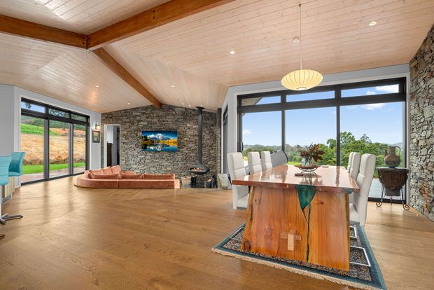 Open-plan living and dining room with a warm timber-lined vaulted ceiling, exposed structural beams, extensive black-framed glazing, and rustic stacked-stone walls. A large sculptural timber dining table anchors the foreground, while a sectional sofa, fireplace, and television occupy the living area. The room overlooks landscaped grounds and has a spacious, bright, nature-connected atmosphere.