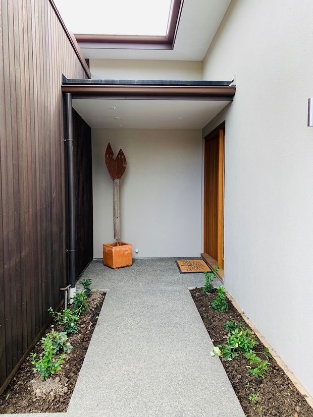 A covered residential entry courtyard with a straight concrete walkway bordered by narrow planted garden beds. The space is enclosed by a dark vertical timber-clad wall on the left and a light rendered wall on the right, with a timber entry door, sheltered ceiling, skylight, and a sculptural outdoor vessel near the entrance.