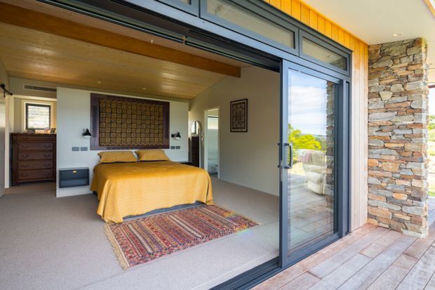 A bright bedroom opening directly onto a timber deck through large sliding glass doors. The room features a low bed with mustard-yellow bedding, patterned wall textile, patterned area rug, dark bedside storage, carpeted flooring, and a timber-lined ceiling with exposed beam. A stacked natural-stone exterior wall and landscaped outlook are visible beyond the glazing.