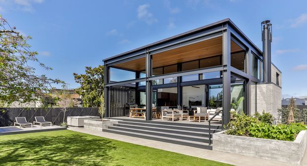 Exterior view of a contemporary two-level outdoor entertaining pavilion or extension with extensive floor-to-ceiling glazing, dark metal framing, a warm timber-lined soffit, and pale masonry side walls. Broad steps lead to a covered terrace furnished for dining and lounging. The landscaped yard includes raised concrete garden beds and a lawn, with a pool or paved relaxation area visible to the left.