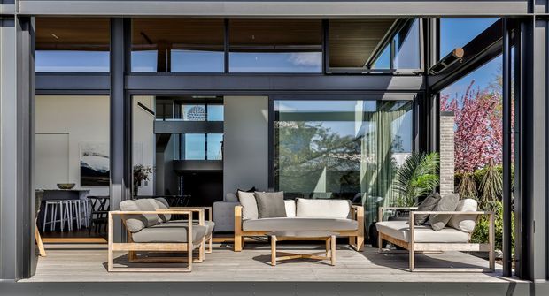 A bright contemporary indoor-outdoor living space framed by extensive dark aluminium glazing and exposed structural elements. The room contains a light timber-framed sofa, armchairs, coffee table, and neutral cushions, with a dining area visible through the opening on the left. Timber ceiling lining, pale flooring, large glass panels, and views toward landscaped planting create a refined connection between the living room and exterior.