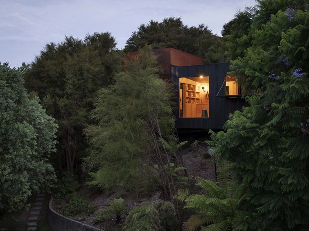 A compact dark-clad architectural cabin or dwelling nestled among dense mature vegetation on a wooded site. Warm interior light glows through large windows and a glazed door, revealing built-in shelving and a small interior space. The elevated structure appears to sit on a steep, landscaped slope, with a narrow stepping-stone path leading through the planting. The image has a moody dusk atmosphere and a strong contrast between the black exterior, rust-toned upper volume, and warm timber-lit interior.