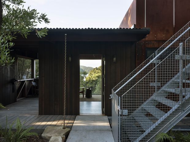 Exterior view of a dark timber-clad building with a corrugated roof, concrete entry path, and an exposed metal stair with wire-mesh balustrades. A chain hangs beside the doorway, and landscaped vegetation and a distant wooded hillside are visible beyond the open passage.