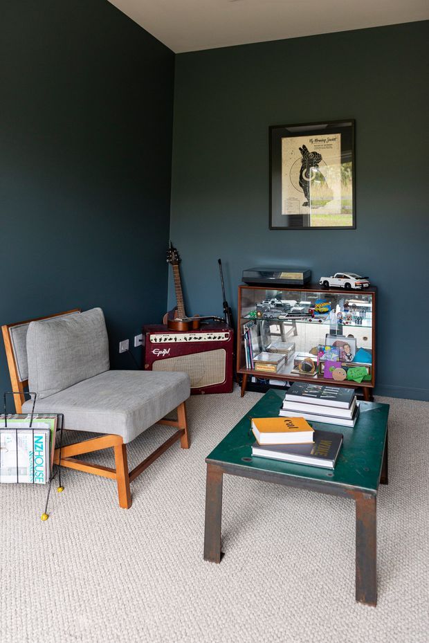 A compact, moody sitting room or study with deep blue-green painted walls, a light grey upholstered lounge chair, a dark green coffee table, a vintage-style guitar amplifier and guitar, and a glass-front display cabinet filled with books and collectibles. Framed artwork hangs above the cabinet, while a light neutral carpet covers the floor.