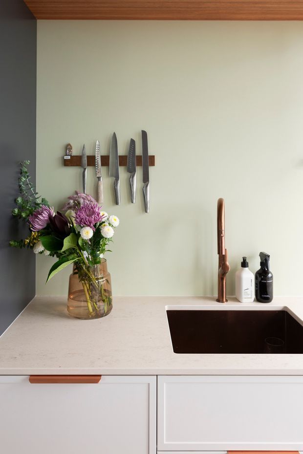 Close-up of a refined kitchen sink zone featuring a pale stone benchtop, deep dark undermount sink, copper-toned gooseneck tap, white cabinetry with timber handles, and a muted sage-green splashback wall. A timber magnetic knife rack, knives, flower arrangement, and soap dispensers add functional and decorative detail. The ceiling or upper cabinetry has a warm timber finish.