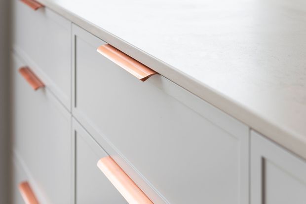 Close-up detail of pale grey shaker-style cabinetry with raised-panel doors and drawers, topped by a light stone or solid-surface benchtop. Distinctive copper-rose tubular handles provide a warm accent against the cool neutral joinery.