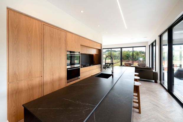 Contemporary open-plan kitchen with extensive warm timber cabinetry, integrated black appliances, and a large dark stone island with a sink and seating. Full-height black-framed glazing opens toward a bright living area and outdoor view. The timber floor is laid in a herringbone pattern, and a linear ceiling light emphasizes the long, refined space.