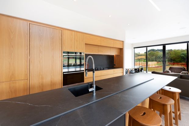 Bright contemporary kitchen with extensive natural timber cabinetry, integrated appliances, a dark stone island benchtop with sink and gooseneck tap, and timber bar stools. Large black-framed glazing opens views toward a landscaped rural setting and adjoining dining/living area.