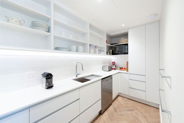 Bright, compact contemporary white kitchen with an L-shaped layout, extensive open shelving, handleless white cabinetry, integrated appliances, a double-bowl sink, and white subway-tile backsplash. Warm herringbone timber flooring adds contrast to the predominantly white scheme.