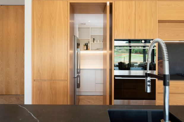 A contemporary kitchen centred on a large dark stone island with an undermount sink and prominent spring-coil gooseneck tap. Floor-to-ceiling light timber cabinetry surrounds integrated appliances, while a concealed pantry with open shelving is visible through the central opening. A window above the benchtop brings in views of greenery, and a timber door is visible at left.