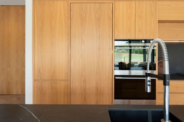 Contemporary kitchen featuring extensive warm timber cabinetry, integrated appliances, a dark stone island benchtop, black undermount sink, and a prominent brushed metal gooseneck tap. A garden is visible through the window behind the cooking area.