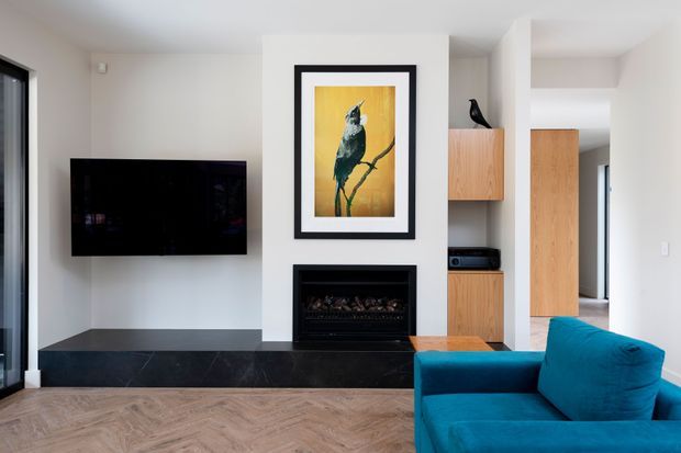 Contemporary living room featuring a wall-mounted black television, black fireplace surround and hearth, framed bird artwork, light timber built-in cabinetry, and a vivid blue upholstered armchair. The room has white walls, dark stone-like fireplace detailing, and a herringbone timber floor.