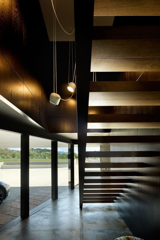 Dark, dramatic interior view of a contemporary entry or stair hall featuring a floating open-riser timber staircase, black structural framing, dark timber wall panelling, polished concrete flooring, full-height glazing, and suspended pendant lights. The glazing opens toward a paved exterior and landscape beyond.