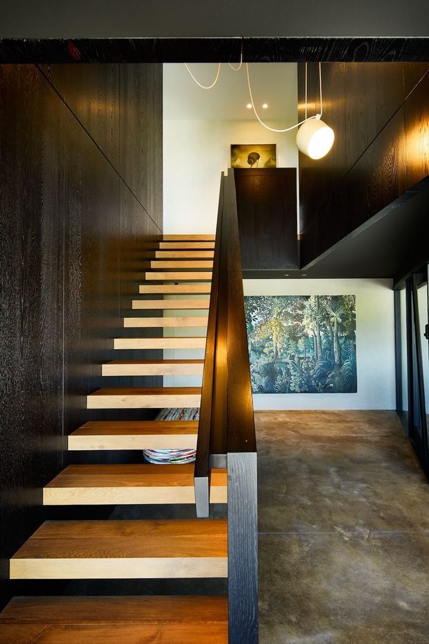 Interior architectural photograph of a modern stairway with floating timber treads, dark timber wall panelling, and a substantial dark timber handrail. The stair rises through a double-height circulation space beside a polished concrete floor, with contemporary pendant lighting, a partially visible upper-level landing, and large artwork on the far wall.