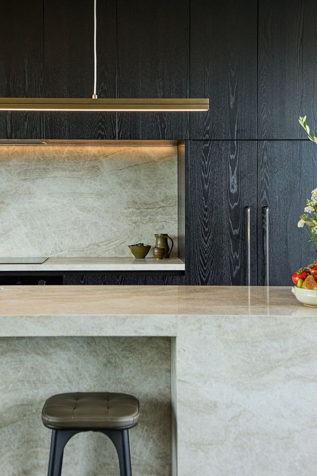 A close-up view of a refined contemporary kitchen featuring dark vertically grained timber cabinetry, a pale veined stone splashback and matching waterfall island. A slim brass pendant light hangs above the island, with a dark upholstered stool beneath the overhang and understated decorative objects on the benchtop.