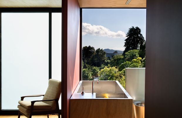 A compact contemporary kitchen or kitchenette positioned beside expansive glazing, with a wood-fronted island or cabinet, stainless-steel sink and faucet, and a small illuminated cooktop or work surface. Warm timber ceiling and joinery contrast with dark window frames and a deep red-brown wall panel. An upholstered lounge chair sits by the window, while lush vegetation and distant hills are visible outside.