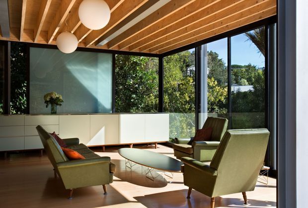 A bright mid-century-inspired living room with large corner glazing overlooking greenery. The room features a warm exposed timber ceiling with beams, dark timber flooring, pale built-in cabinetry, an opaque glass feature panel, sculptural pendant lights, and a grouping of vintage-style lounge chairs around a low oval coffee table.