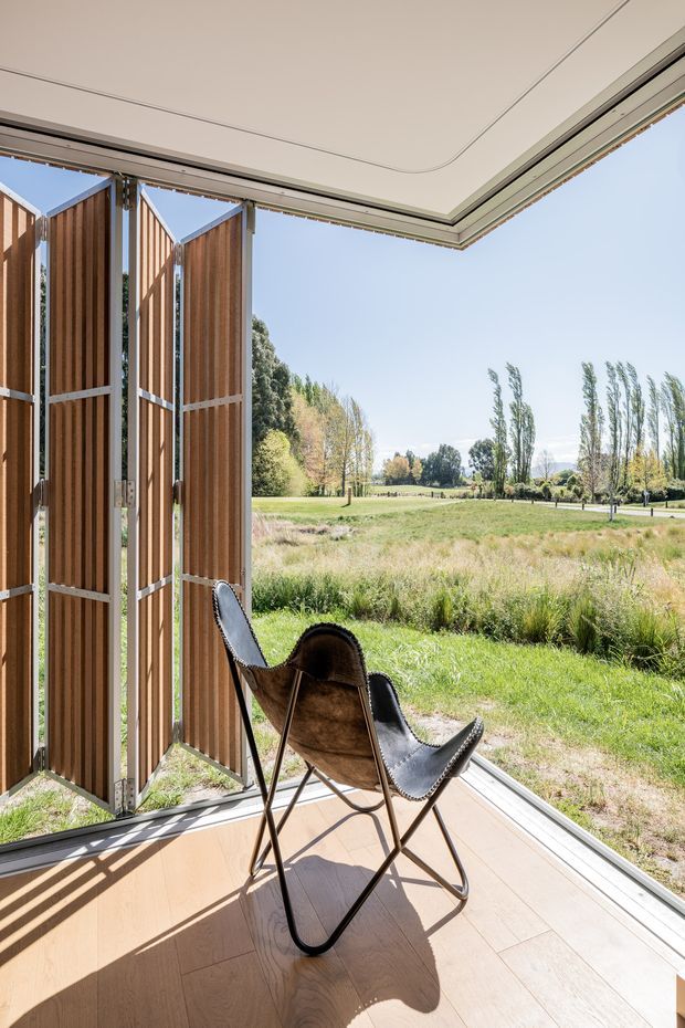 A bright, minimalist indoor-outdoor sitting area with fully opened timber louvred folding screens and extensive corner glazing overlooking a landscaped rural garden. A single leather sling chair sits on a pale timber floor, with a clean white ceiling and slim dark window framing. The space feels airy and strongly connected to the surrounding landscape.