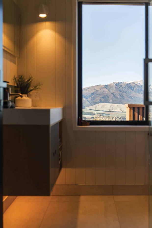 A warmly lit compact interior with vertical timber wall panelling, a dark built-in cabinet or kitchenette with a light benchtop, and a large black-framed window opening to a clear mountain landscape. A small plant and pendant light add warmth, while the view and timber detailing give the space a refined alpine-cabin character.