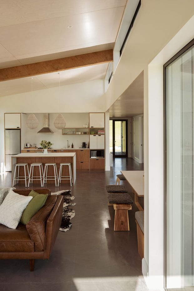 A bright open-plan interior combining a kitchen, dining nook and living area. The kitchen features warm timber cabinetry, a pale benchtop, open shelving, integrated appliances and a compact island with four stools. A large brown leather sofa occupies the foreground, while a timber dining bench with upholstered stools runs alongside expansive glazing. The room has a high sloped ceiling with exposed timber beam, polished dark flooring, pendant lights and abundant natural light.