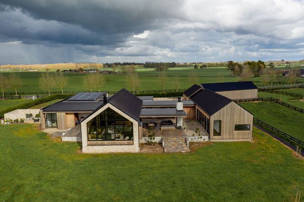Aerial view of a contemporary rural homestead composed of several connected gabled volumes. The house features dark pitched roofs, natural timber-clad walls, large glazed openings, a stone-faced central living pavilion, and a paved courtyard, set within expansive green farmland and fenced paddocks under dramatic overcast skies.