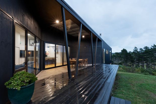 Exterior view of a dark-clad modern single-storey residence with a long covered terrace and wet timber deck. Floor-to-ceiling glazing reveals a warm, open-plan interior, while slender angled posts support the roof canopy. The house sits beside a wooded landscape on a sloping site.