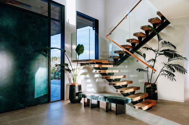 Contemporary residential stairway with open timber treads, dark structural supports, and a clear glass balustrade with timber handrail. The stair rises beside tall black-framed glazing and a dark green feature wall, with indoor plants and a patterned upholstered bench beneath. The space is bright and refined, with polished concrete or large-format grey flooring and a strong indoor–outdoor connection visible through the glazing.
