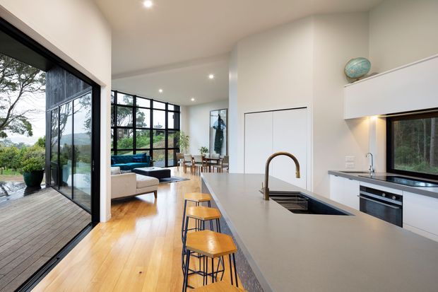 Open-plan contemporary kitchen and living area with a long grey island and integrated sink, timber bar stools, white flat-panel cabinetry, dark benchtops, and timber flooring. Full-height black-framed glazing connects the interior to a timber deck and leafy landscape, while the living and dining areas extend beyond the kitchen.
