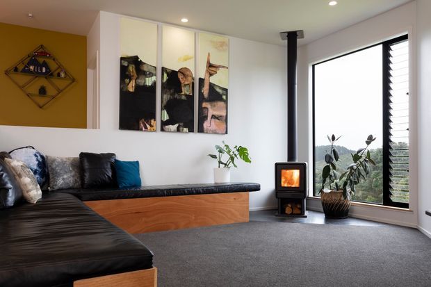 Contemporary living room with a large black sectional sofa, timber-faced built-in bench seating, grey carpet, white walls, and a freestanding black wood-burning stove. A tall louvred window provides views outside, while colourful wall art and indoor plants add visual interest.