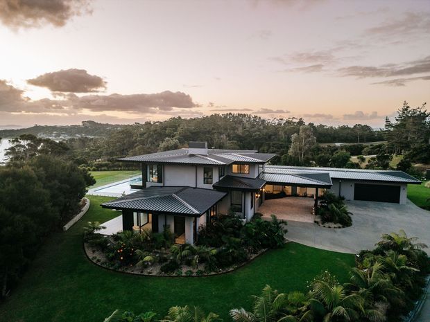 Aerial view of a large contemporary two-storey residence set within expansive landscaped grounds. The home has dark metal hipped and skillion roof forms, light rendered walls, extensive glazing, covered outdoor areas, and a separate garage or outbuilding. A swimming pool is visible beside the house, with mature trees and manicured lawns surrounding the property under a soft evening sky.