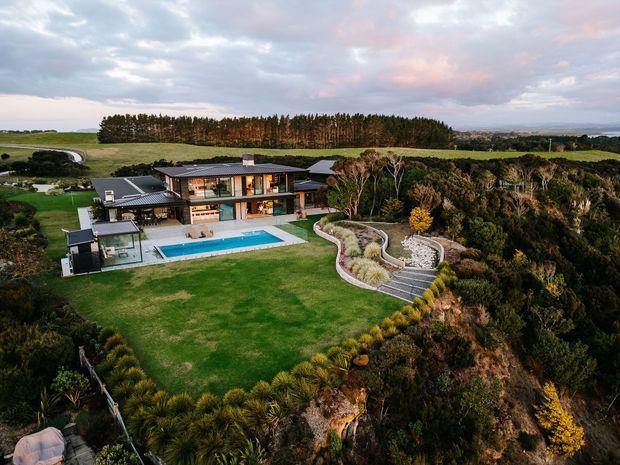 Aerial view of a large contemporary multi-level residence set on a landscaped coastal or rural site. The home has dark low-pitched roof forms, extensive glazing, timber-toned exterior elements, a large swimming pool, paved terraces, and sculpted native-style planting around a dramatic sloping or elevated landscape. A tree-lined rural outlook and distant water or hills are visible beyond the property.