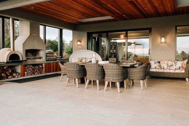 A spacious, open-plan outdoor entertaining area with a dining table, woven dining chairs, a built-in fireplace and pizza oven, and a cushioned lounge seat. Large glazed openings connect the space to the surrounding landscape, while a rich timber-lined ceiling adds warmth above a neutral tiled floor.