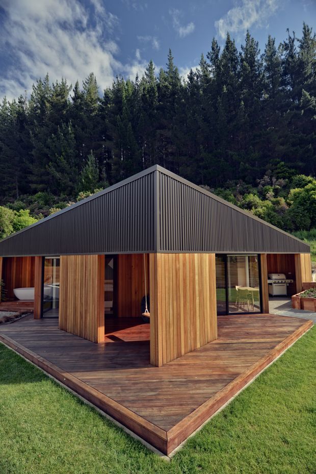 A compact single-storey pavilion-style house set against a dense forested hillside. The building has a steep gabled roof clad in dark vertical corrugated metal, warm vertical timber wall cladding, expansive black-framed glazing, and a broad timber deck wrapping around the front. A partially visible outdoor kitchen or barbecue is located at the right, with a bathtub visible through the left glazing.
