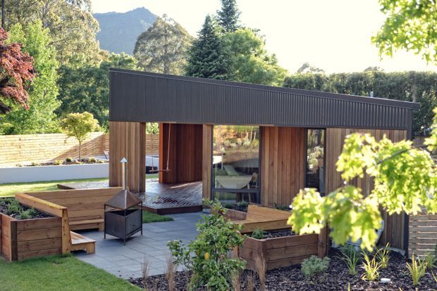 A contemporary single-storey garden pavilion or outdoor living structure set within landscaped grounds. The building has dark vertically ribbed cladding, warm timber-lined walls, extensive glazing, and a strong asymmetrical sloping roofline. A paved courtyard with raised timber planter beds, built-in bench seating, and a freestanding outdoor fireplace or fire pit occupies the foreground, with mature vegetation and mountainous terrain beyond.