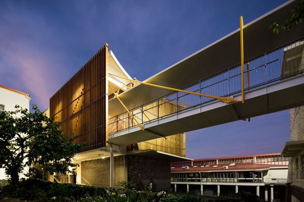 Twilight exterior view of a contemporary multi-level building with a prominent elevated pedestrian bridge or balcony, dark metal railings, warm timber vertical screening, exposed structural elements, and a sharply angled roof form. The complex sits in an urban setting with adjacent buildings and landscaped planting visible at ground level.