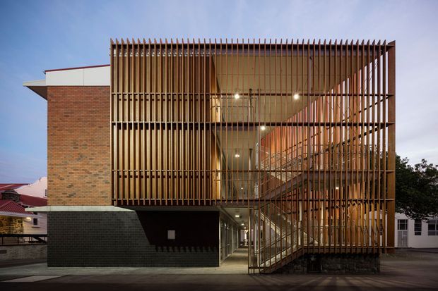 Exterior architectural photograph of a multi-storey brick building with a prominent open stairwell and entry atrium screened by dense vertical timber fins. The facade combines exposed brick, dark masonry at the lower level, projecting roof planes, warm interior lighting, and a visible external stair, creating a strongly articulated contemporary institutional or commercial frontage.