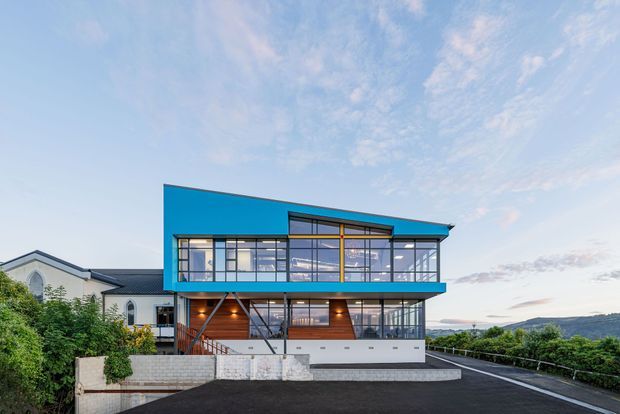 Contemporary multi-storey residence with a bold turquoise-blue upper facade, extensive floor-to-ceiling glazing, timber-toned lower-level elements, and a dark low-pitched roof. The home sits on a sloping site with a paved driveway, retaining walls, landscaped planting, and distant coastal or hilly views.